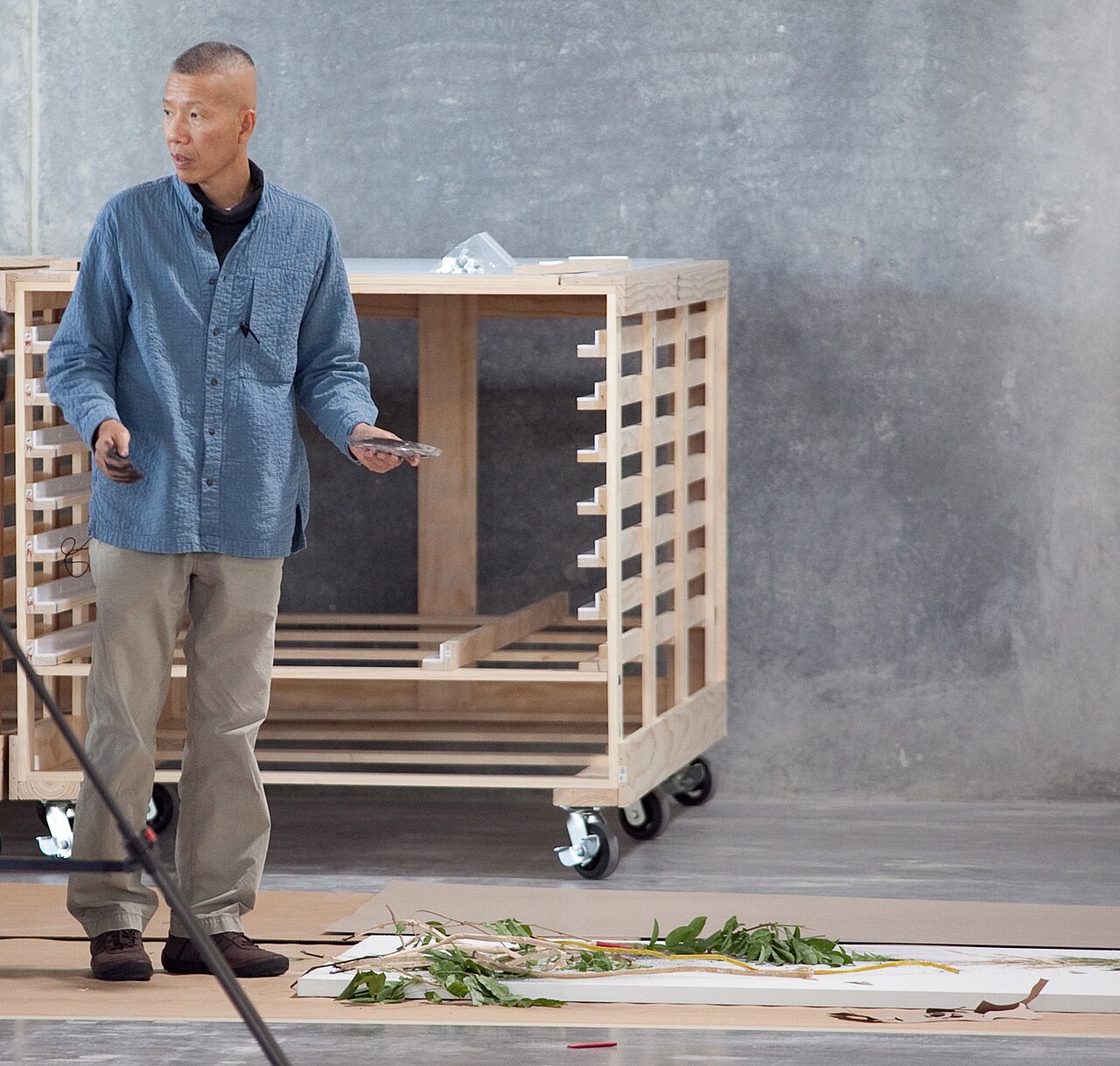 Cai Guo-Qiang standing in his studio beside a large wooden crate, with plant materials laid out on canvas on the floor
