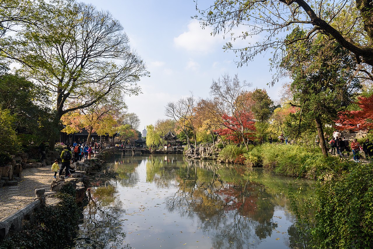 An autumn view of the Humble Administrator's Garden in Suzhou with a tranquil pond reflecting traditional pavilions and colorful foliage