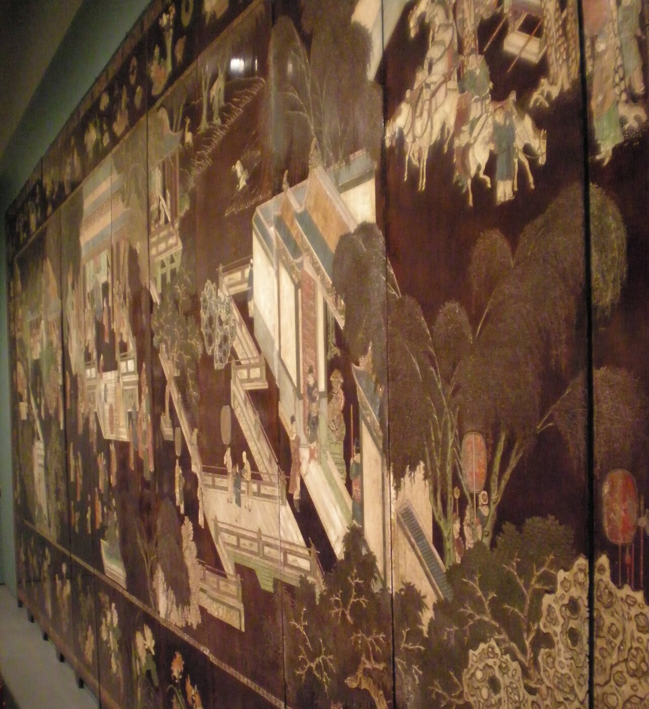 A large twelve-panel Coromandel lacquer screen showing a detailed palace garden scene with figures and architecture in polychrome on dark background