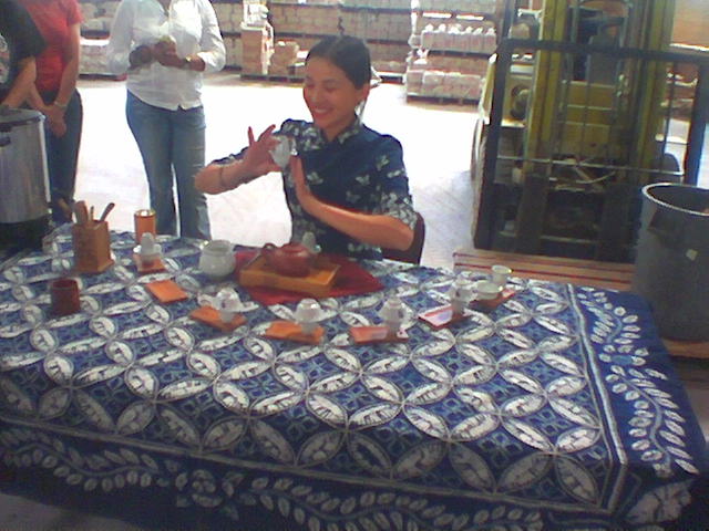 Woman in a traditional qipao performing a Chinese tea ceremony with multiple cups on a blue batik tablecloth
