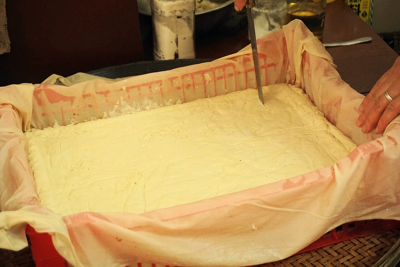 Fresh tofu being sliced from a traditional cloth-lined pressing mold