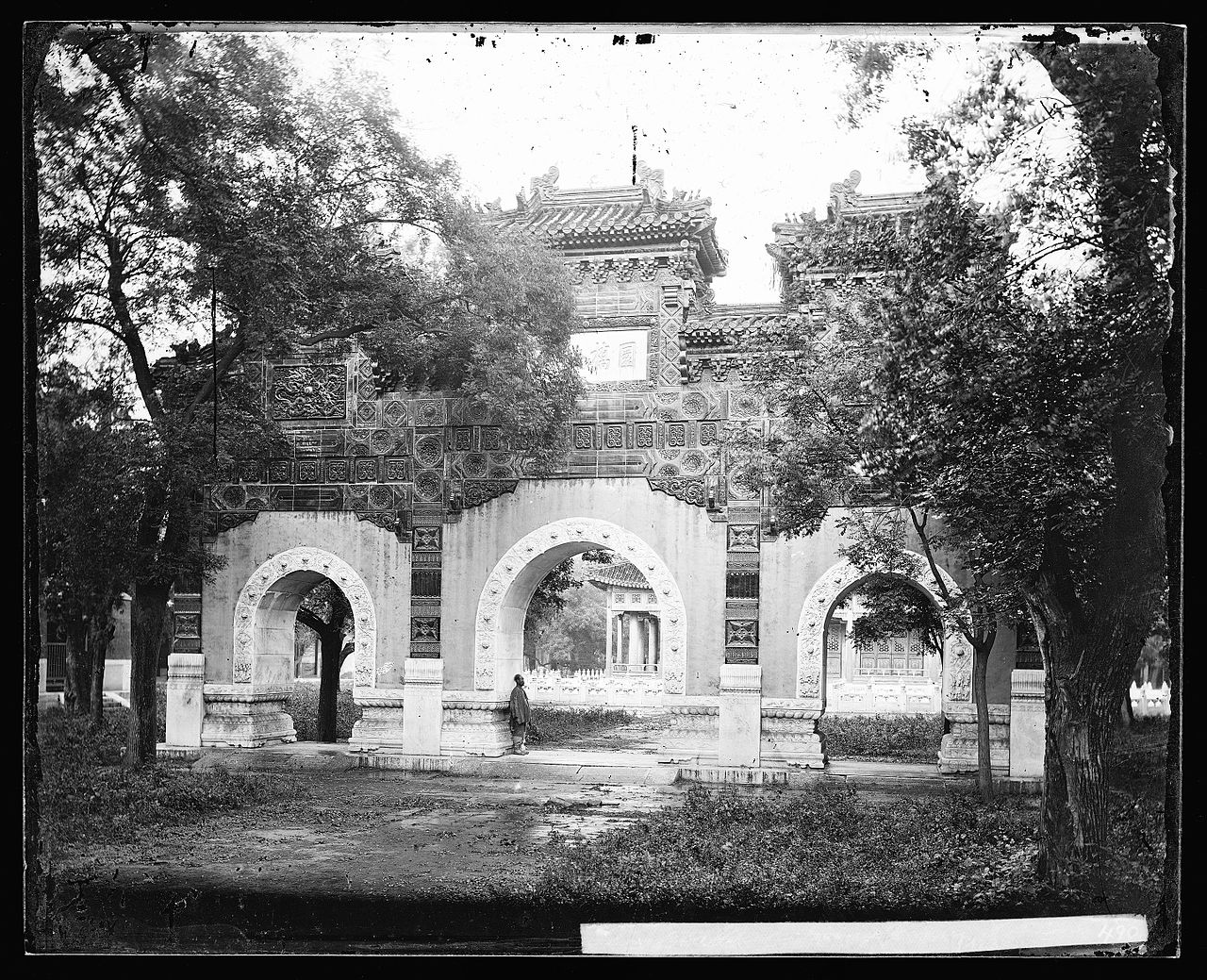 Historic black and white photograph of the ornate triple archway at the Guozijian (Imperial Academy) Hall of Classics in Beijing, with a solitary figure standing beneath the central arch