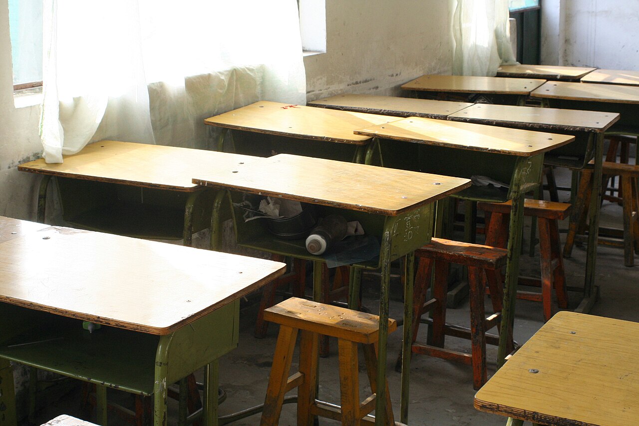 Empty Beijing school classroom with rows of green metal-framed wooden desks and stools, sunlight streaming through curtained windows