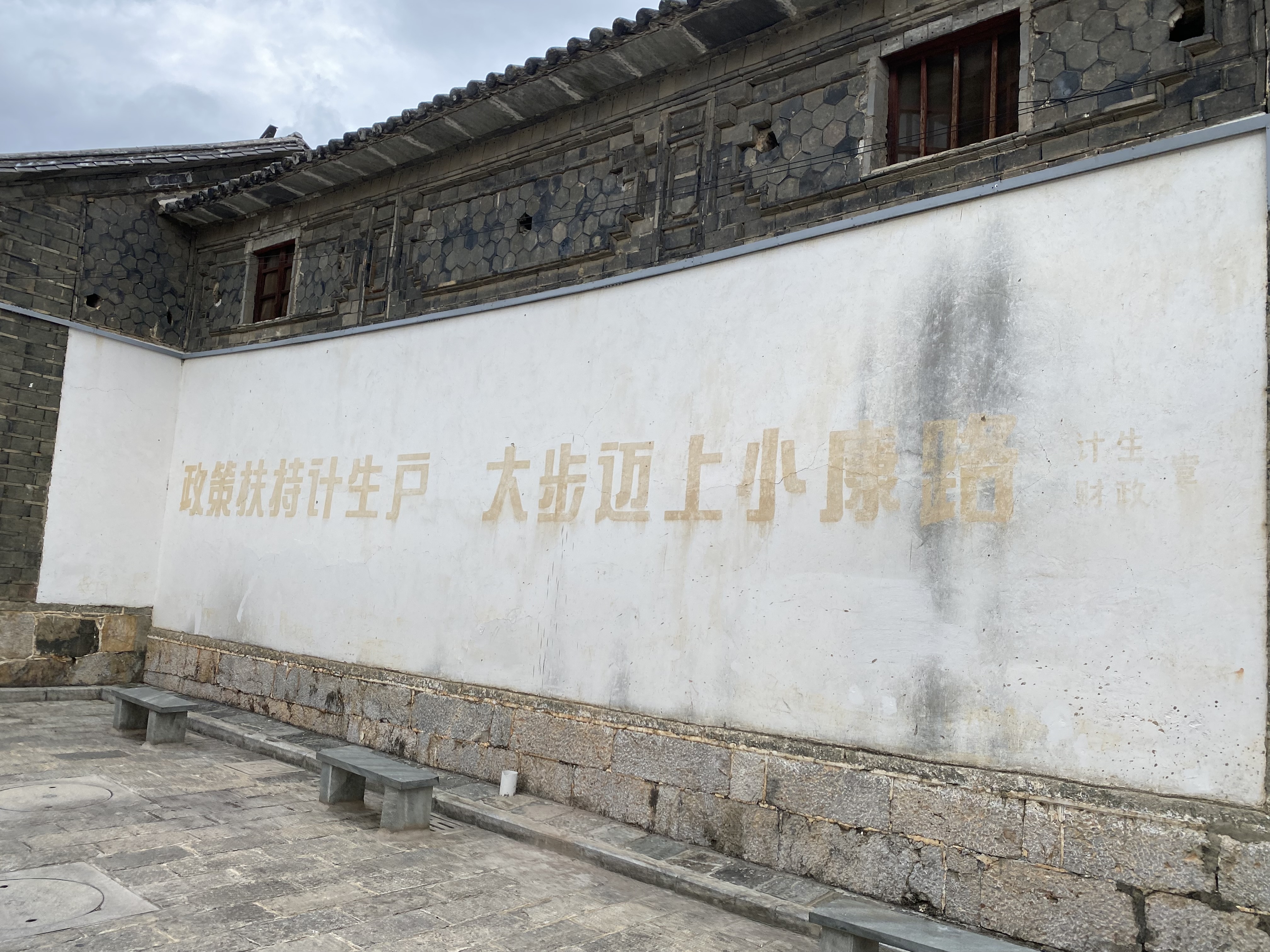 Faded yellow Chinese family planning slogan painted on a white wall of a traditional stone building in Dali City, China