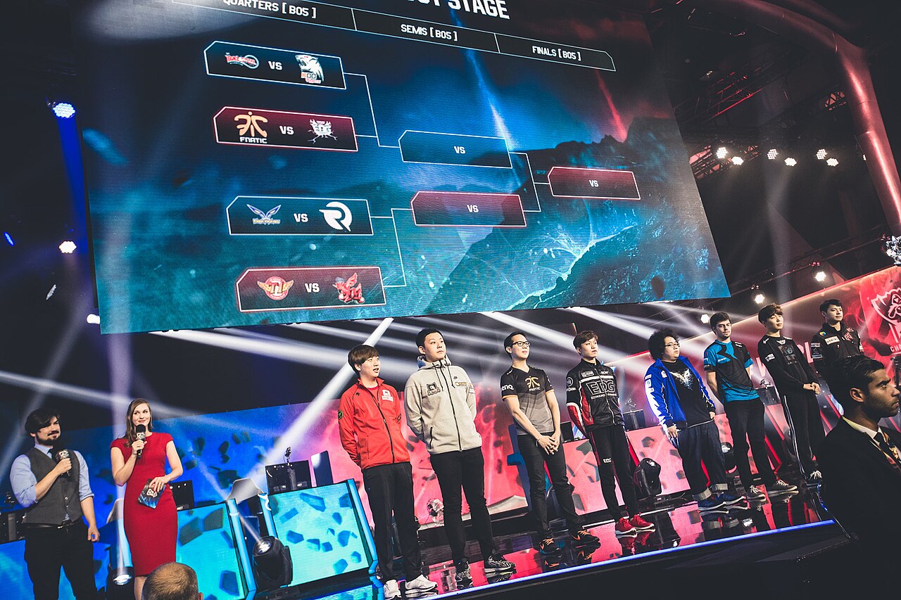 Professional esports players from multiple teams standing on stage at the 2015 League of Legends World Championship, with a tournament bracket displayed on a large screen behind them