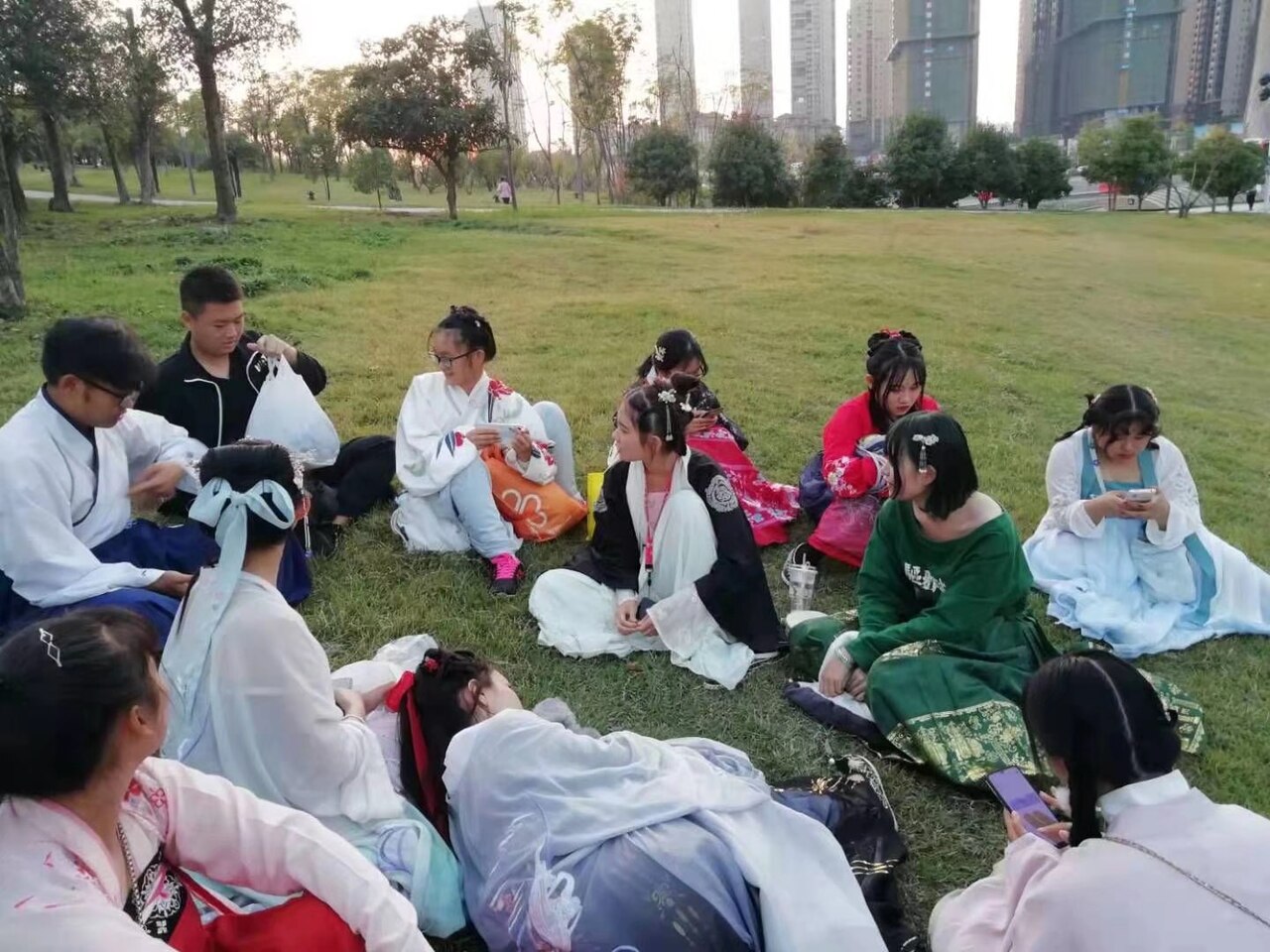 Group of young Chinese students wearing colorful hanfu traditional clothing sitting together on grass in a park, with city buildings visible in the background